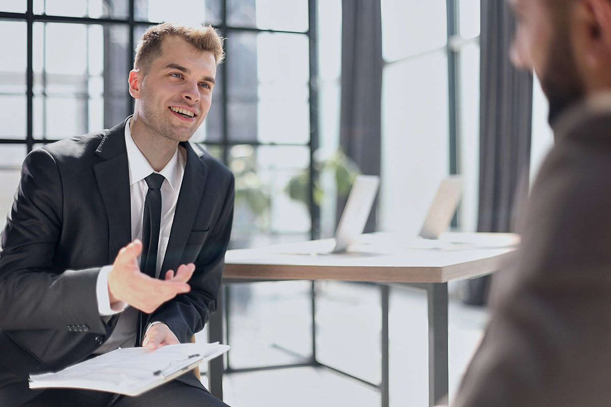 A young man in a suit and tie is engaged in a conversation, smiling and gesturing while holding a clipboard, sitting across from another person in a well-lit, modern office space.