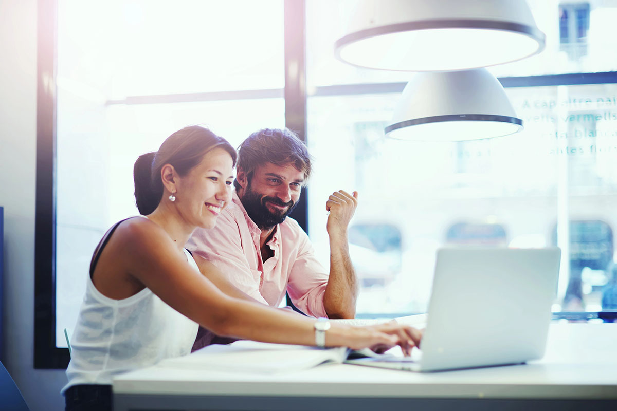 A woman and a man are sitting at a table, smiling and engaging with a laptop in a bright, modern setting. The woman is leaning forward, actively typing, while the man looks pleased and gestures with his hand.