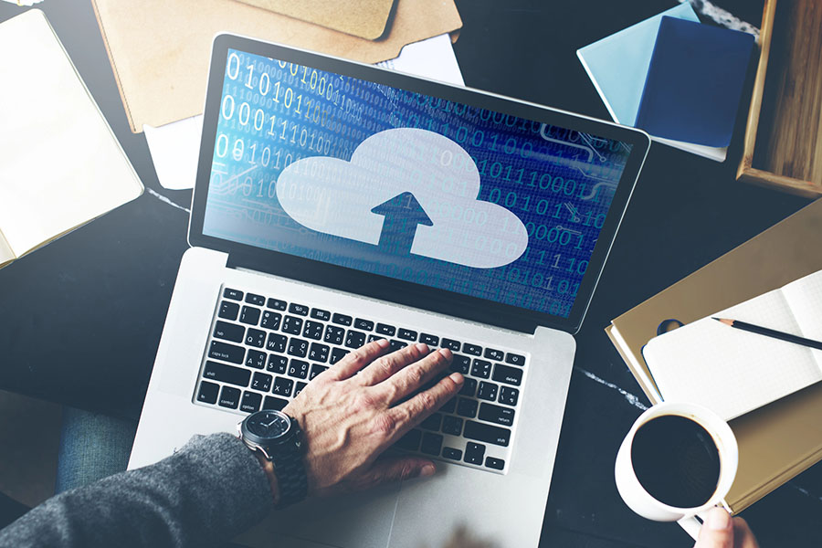 A close-up view of a laptop displaying a cloud upload symbol against a background of binary code, with a hand typing on the keyboard and a coffee cup nearby.