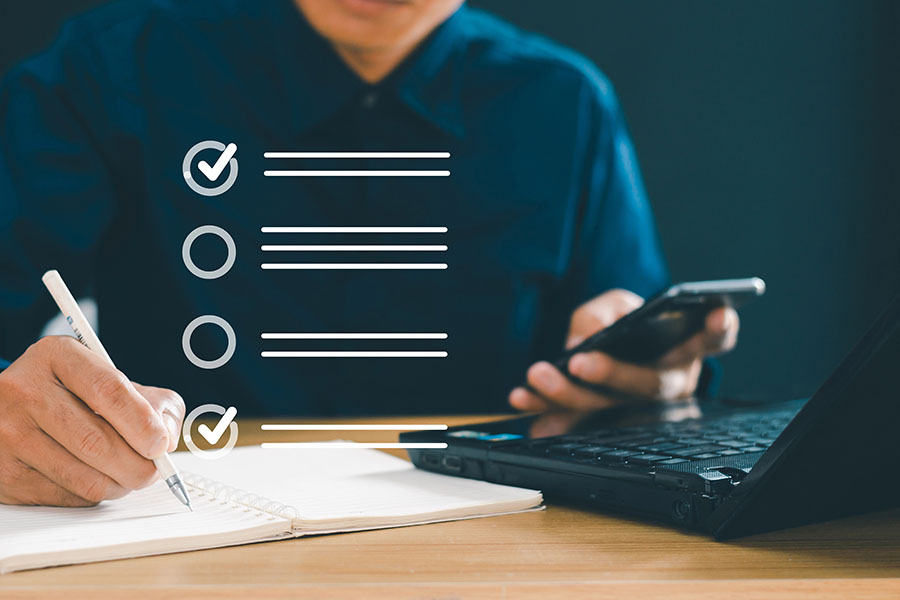A person sits at a wooden table, writing in a notebook with a pen in one hand while holding a smartphone in the other. In the background, a laptop is visible, and a checklist graphic with checkmarks and empty lines appears above the notebook.