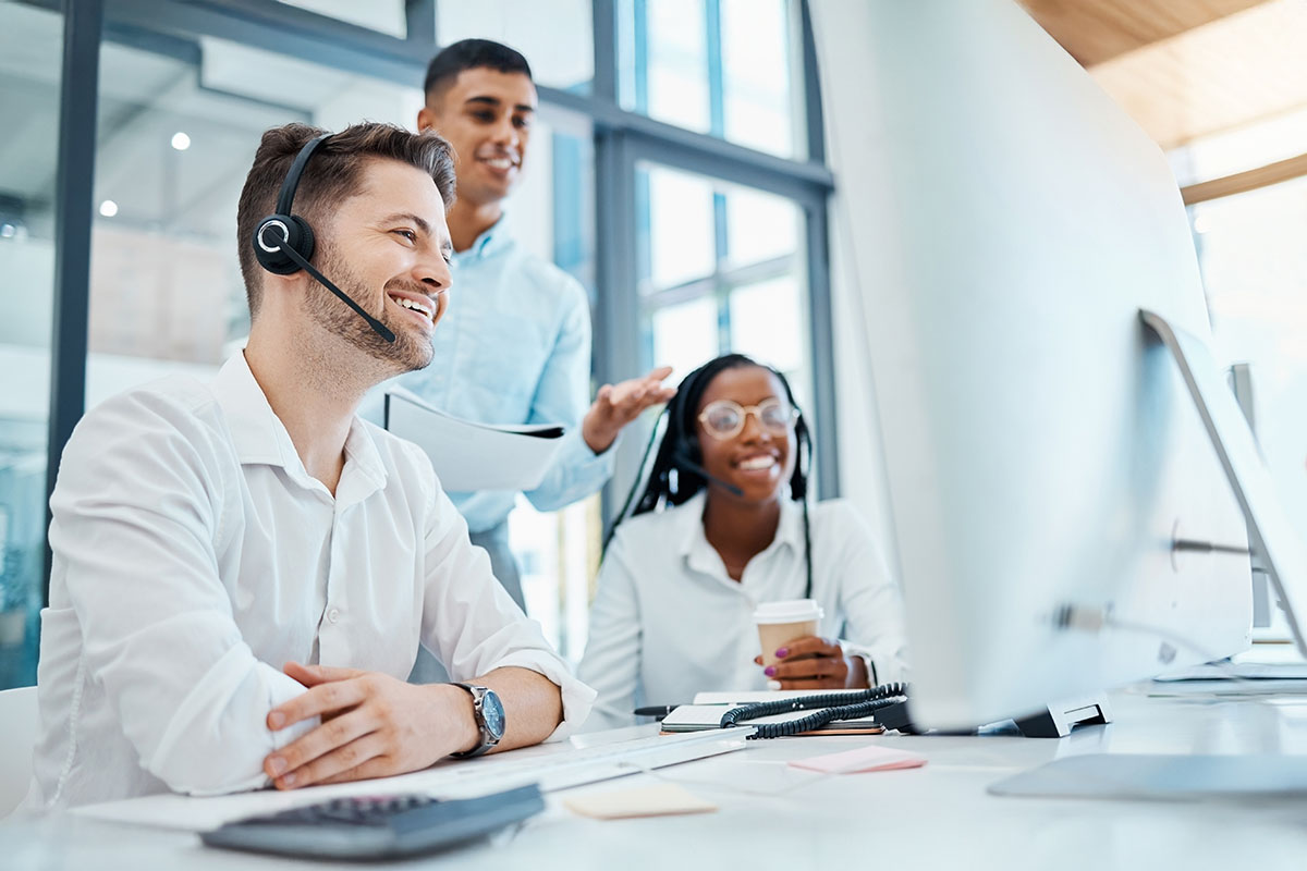 Three professionals in an office setting: a man wearing a headset smiles at a computer, while a woman with glasses holds a coffee cup and a man in a blue shirt gestures while holding a document.