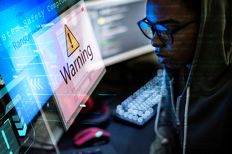 A person wearing a hoodie and glasses looks intently at a computer screen displaying a warning message about ransomware, surrounded by digital code and graphics.