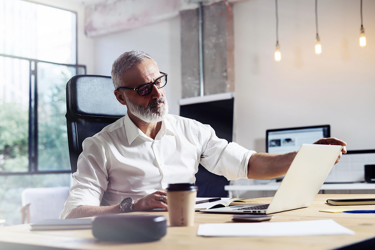 A bearded older man in glasses sits at a desk in a modern office, focused on his laptop. He wears a white shirt and has a coffee cup and notebook on the table. Natural light streams through large windows in the background.