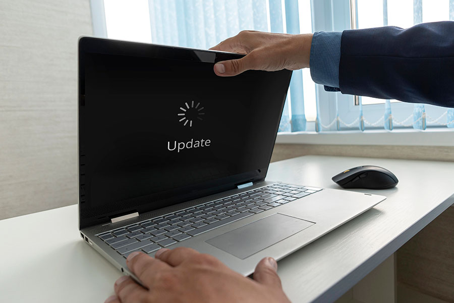 A person in a suit is about to close a laptop with an "Update" loading screen displayed on the screen, while a computer mouse is positioned nearby on a white desk with a window in the background.