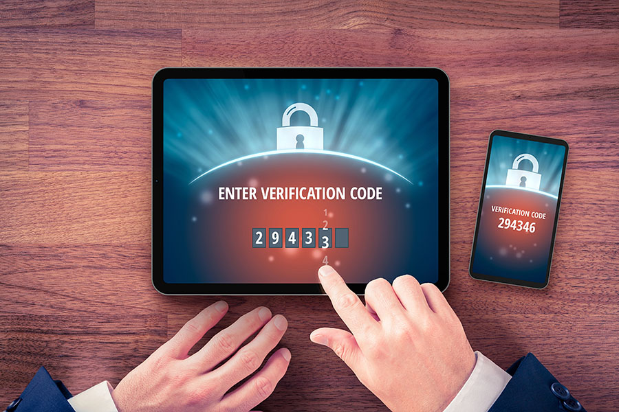 A person’s hands are shown interacting with a tablet displaying a prompt to "ENTER VERIFICATION CODE" alongside a smartphone showing the code "294346." Both devices have a padlock graphic in the background.