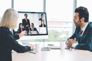 A woman in a business suit gestures during a video conference, engaging with five participants displayed on a large monitor. Two mugs and a notebook are on the table in front of her, while a man in a suit observes her from the side.