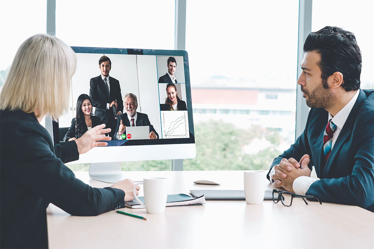 A woman in a business suit gestures during a video conference, engaging with five participants displayed on a large monitor. Two mugs and a notebook are on the table in front of her, while a man in a suit observes her from the side.