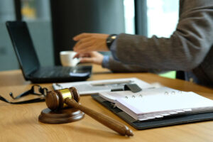 A man wearing a suit is sitting at a computer desk with a binder, gavel, and laptop on the desk.