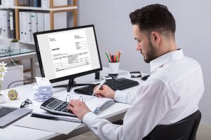 A man in a white shirt is seated at a desk, focused on his work. He is using a calculator with one hand while writing on a document with the other. A computer monitor displays an invoice. The desk is cluttered with papers, a laptop, and office supplies, including a cup of pens and a small plant.