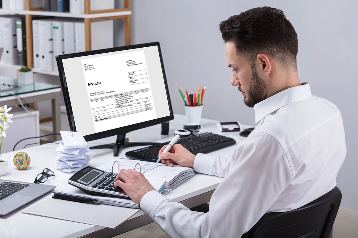 A man in a white shirt is seated at a desk, focused on his work. He is using a calculator with one hand while writing on a document with the other. A computer monitor displays an invoice. The desk is cluttered with papers, a laptop, and office supplies, including a cup of pens and a small plant.