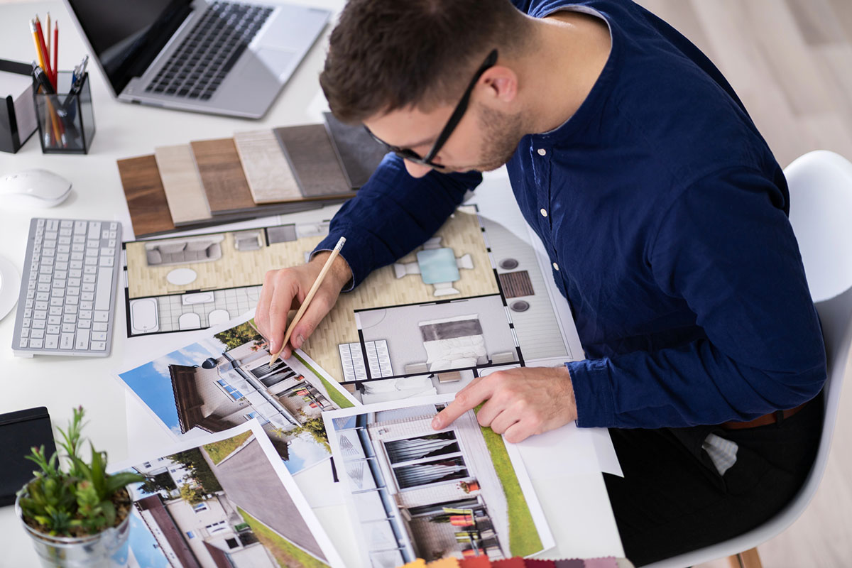 A man with glasses is seated at a desk, reviewing architectural plans and photographs of houses. He uses a pencil to make notes on the plans, surrounded by samples of flooring materials, a laptop, and a keyboard. A small plant is visible in the foreground.