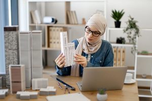 A woman wearing a hijab and glasses sits at a desk, examining a detailed building model while surrounded by several other architecture models, a laptop, and drafting tools in a modern workspace.