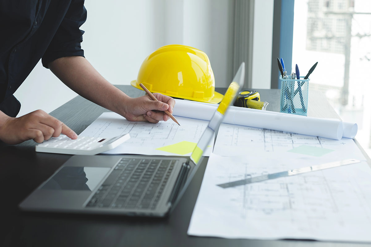 A close-up of a person's hand using a calculator while writing on architectural plans, with a laptop, yellow hard hat, and stationery visible on a desk.
