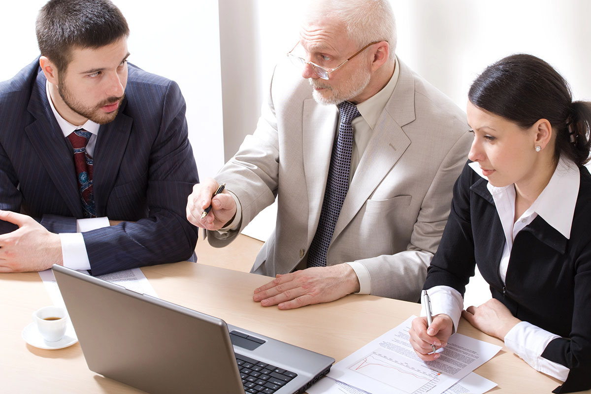 Three business professionals are seated at a table discussing work. The man in a dark suit appears focused, while an older man in a light suit gestures with a pen, pointing at a laptop and documents. A woman in a black blazer listens attentively, taking notes. A small cup of coffee is visible on the table.