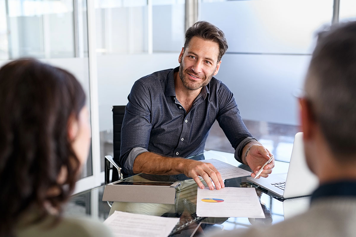 A man in a dark shirt sits at a conference table, smiling and engaging in conversation with two individuals whose faces are out of focus. Papers and a laptop are on the table.