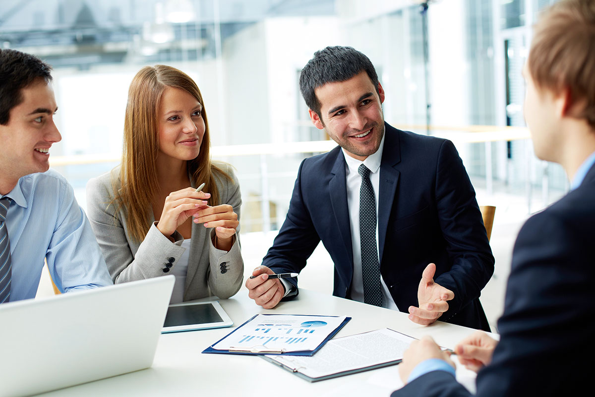 A group of four professionals sitting around a table in a modern office, engaged in discussion. Two men and two women are dressed in business attire, with laptops and documents on the table, conveying a collaborative work environment.