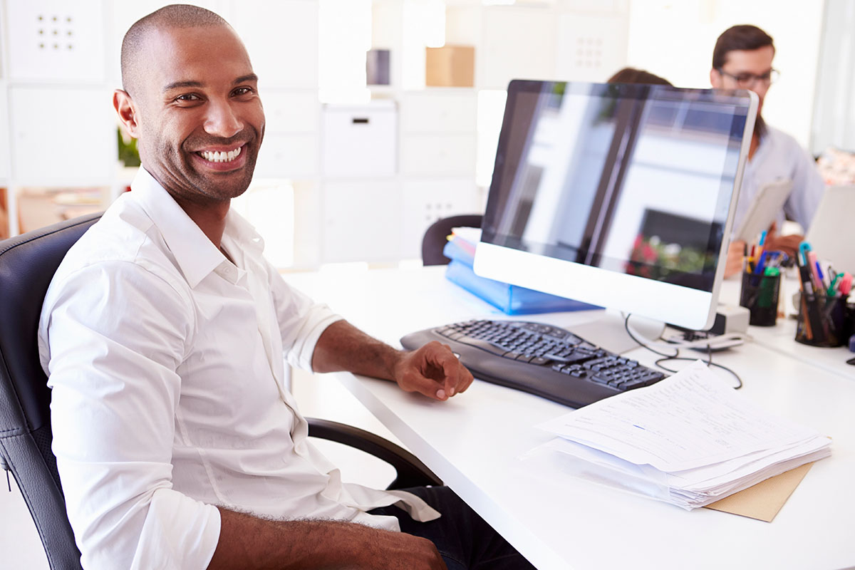 A smiling man in a white shirt sits at a desk with a computer, keyboard, and paperwork, in a bright office environment with colleagues in the background.