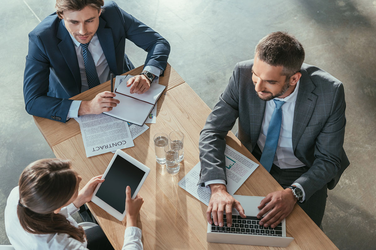 Three professionals in business attire are seated around a wooden table, discussing documents and analyzing data. One person is using a laptop, another is holding a tablet, and the third is taking notes on a notepad. Glasses of water and various papers are spread across the table.