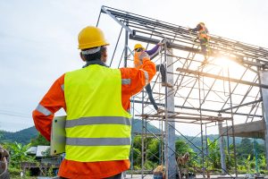 A construction worker in a hard hat and reflective vest watches as two other workers climb a scaffolding to work on a building frame, with a mountainous landscape in the background.