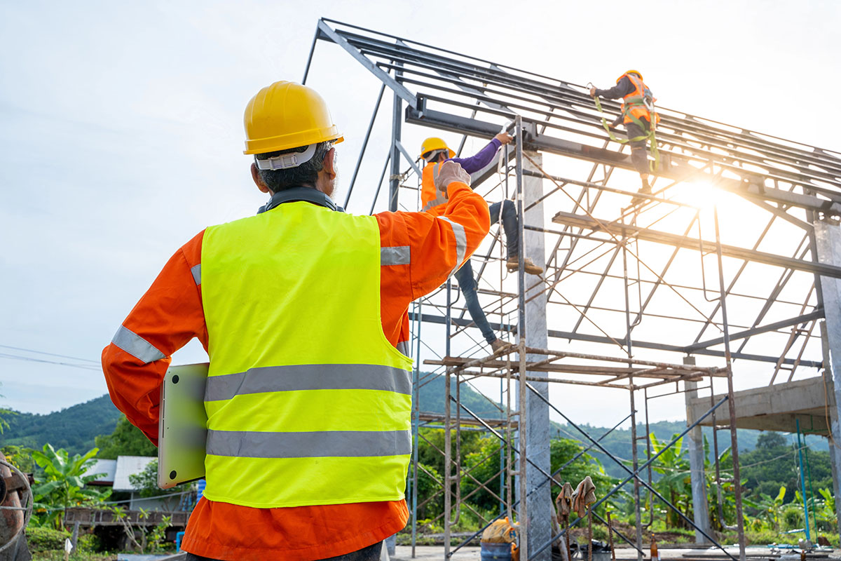 A construction worker in a hard hat and reflective vest watches as two other workers climb a scaffolding to work on a building frame, with a mountainous landscape in the background.
