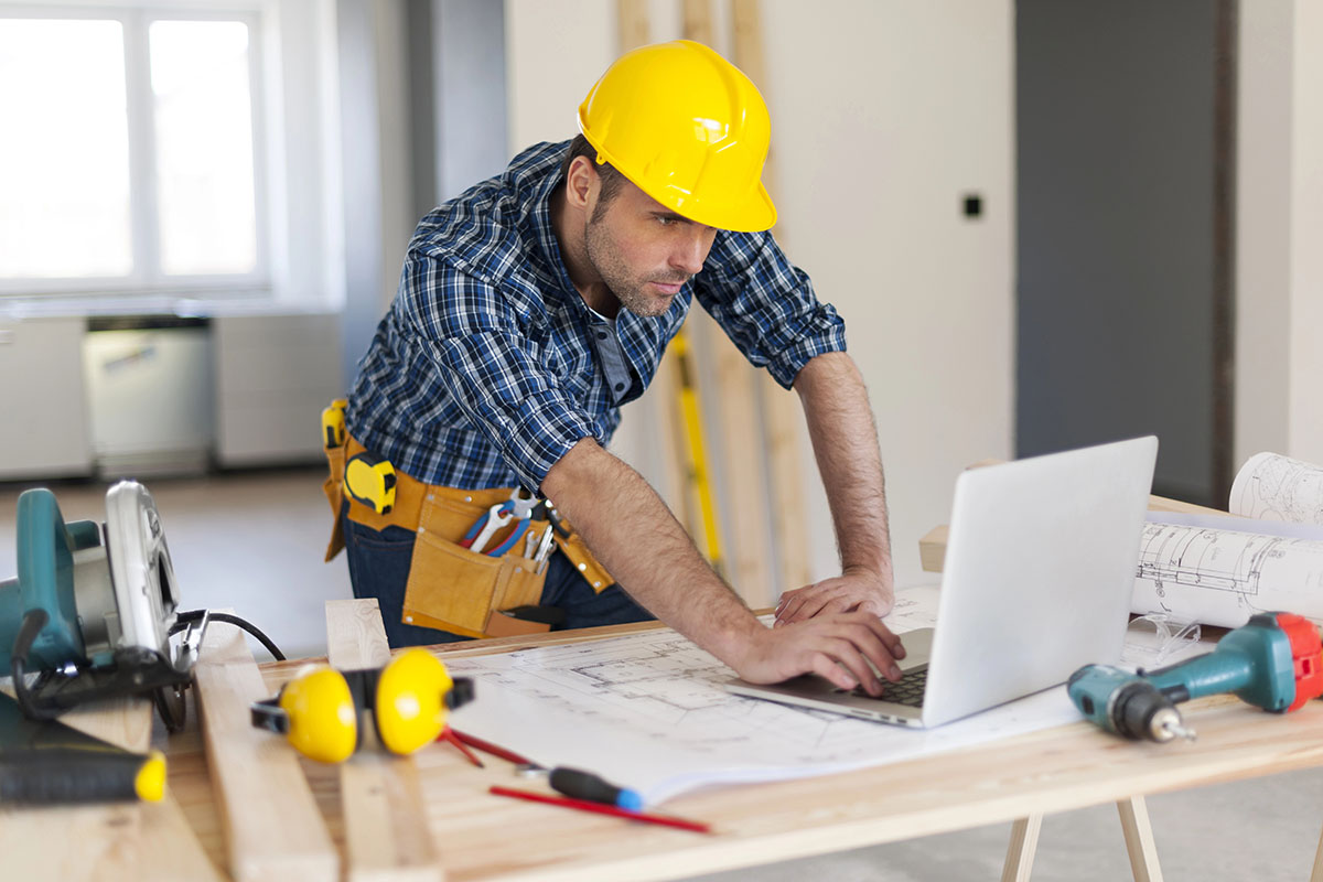 A construction worker in a yellow hard hat is focused on a laptop while leaning over a table filled with blueprints and tools, including a circular saw, a drill, and measuring equipment, in an unfinished room.