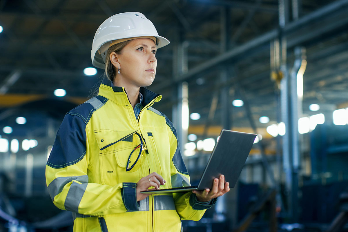 A woman in a bright yellow safety jacket and white hard hat is standing in an industrial setting, holding a laptop and gazing thoughtfully into the distance.