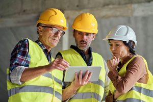 Three construction workers, two wearing yellow hard hats and vests and one wearing a white hard hat and a brown shirt, are gathered around a tablet, discussing plans on a construction site.