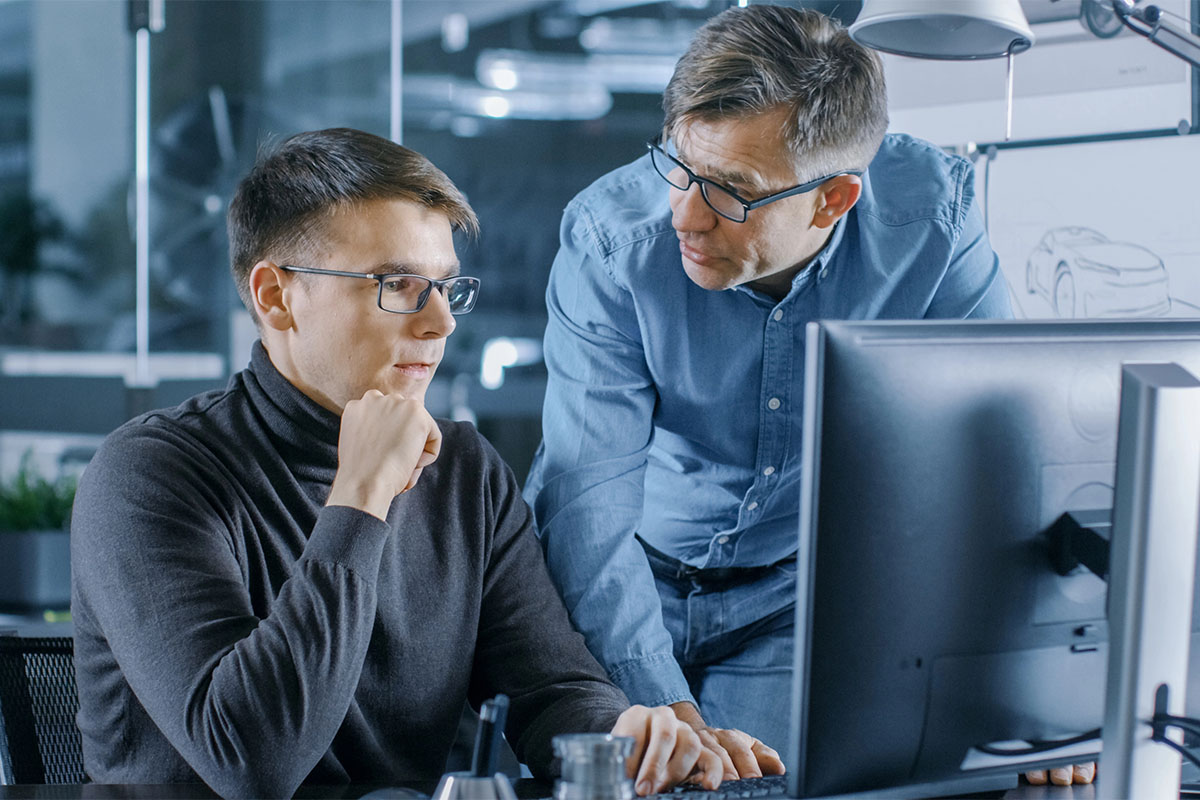 A young man in a black turtleneck sweater sits at a desk, thoughtfully resting his chin on his hand while looking at a computer screen. Beside him, an older man in a blue button-up shirt leans over, engaging in discussion. The background features modern office elements and a design sketch of a car.