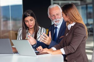 Three business professionals are engaged in a discussion around a laptop at a table. The first woman, with long dark hair and a striped blouse, is speaking while showing concern. The older man with a beard and glasses is listening intently, gesturing with his hands. The second woman, with long light brown hair and a brown suit, is also focused on the conversation. The setting appears to be a modern office with large windows.