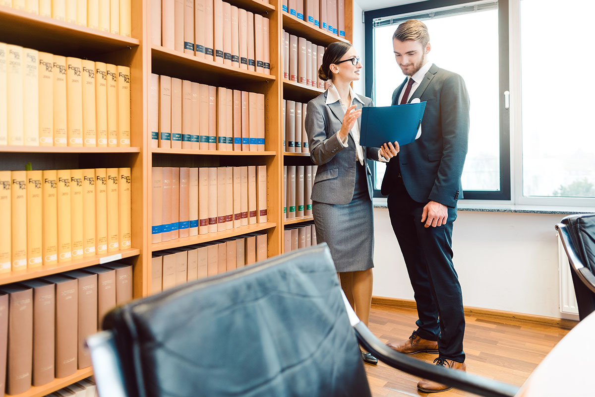 A professional man and woman stand in an office library, discussing a document. Bookshelves filled with law books are in the background, and a large window lets in natural light. The woman is wearing a grey business suit and glasses, while the man is in a dark suit and tie.