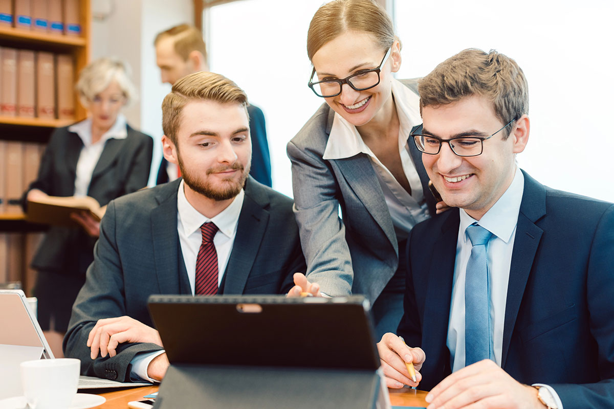 A group of four professionals is engaged in a collaborative meeting. Two men in business attire are focused on a laptop, while a woman in a suit points at the screen, smiling. In the background, another woman is holding a book, and a man is standing nearby. The setting is a modern office with shelves of books.