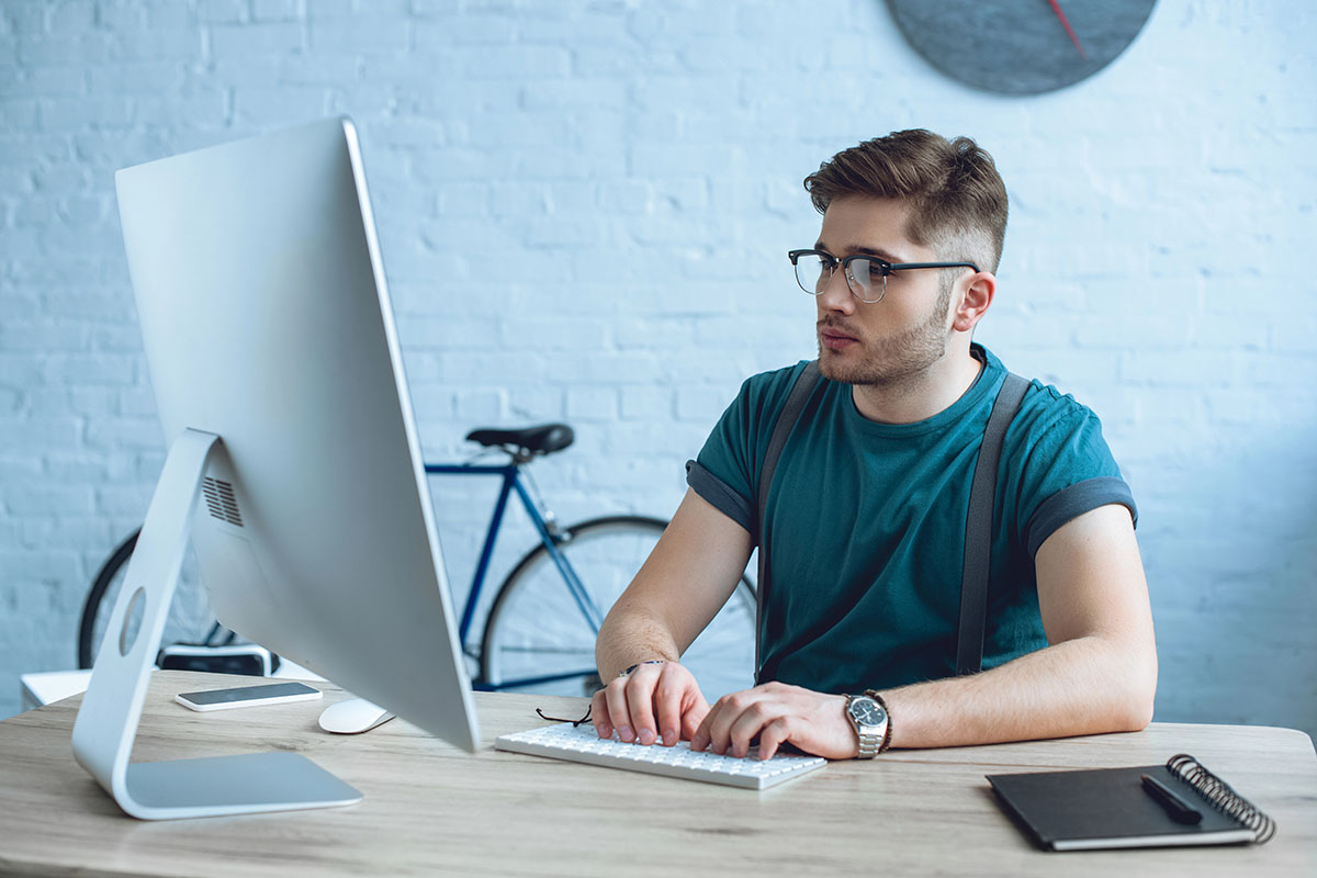 A young man with glasses sits at a wooden desk, focused on a computer screen. He wears a teal t-shirt and has suspenders. In the background, a blue bicycle leans against the wall, and a round clock is visible on the wall.