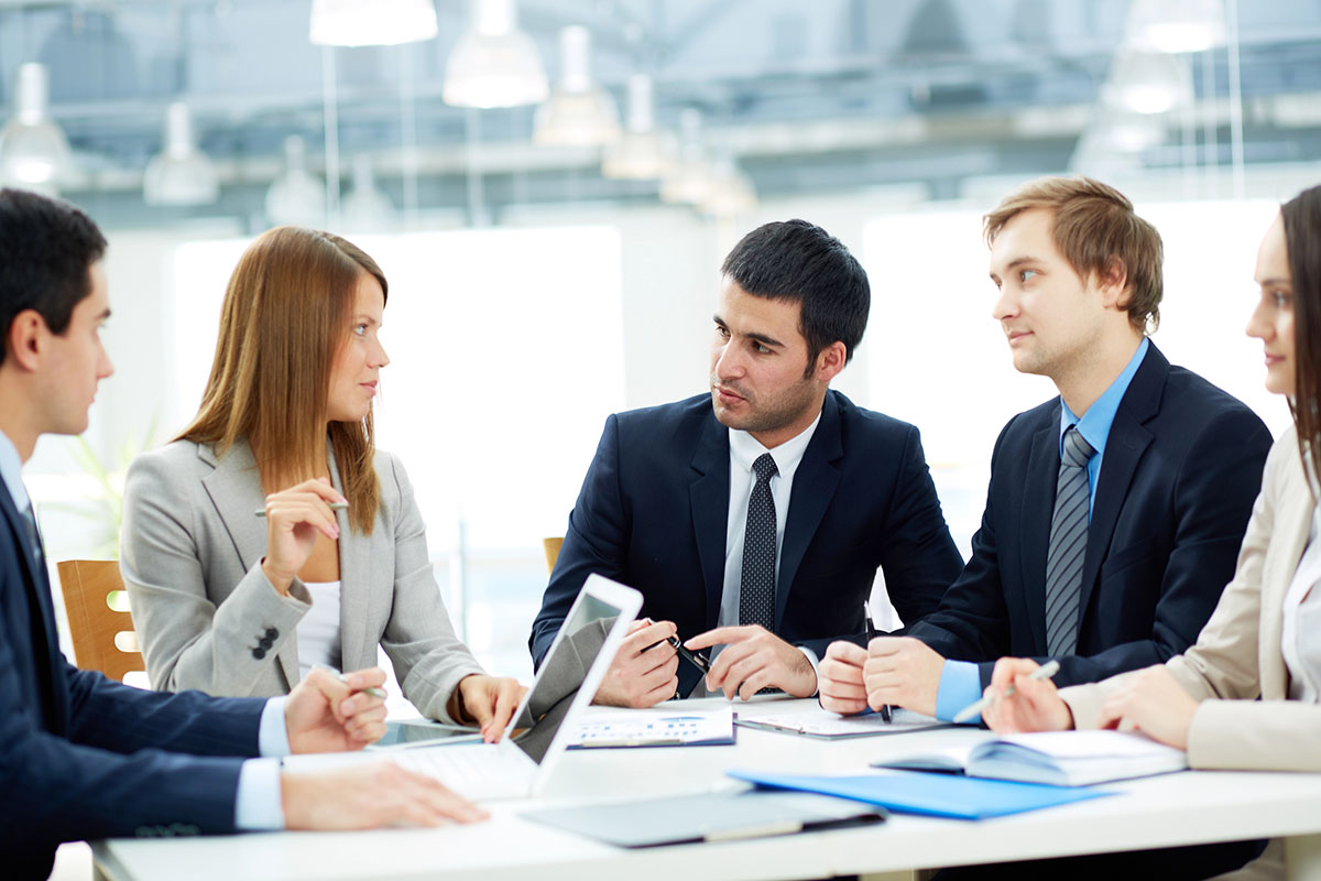 A group of five professionals engaged in discussion around a conference table, with laptops and notepads visible. They are dressed in formal business attire, displaying a mix of expressions as they communicate. Natural light filters in from a nearby window.