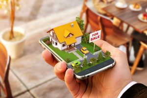 A hand holding a smartphone displays a 3D model of a house with an orange roof, a garden, and two small cars parked in the driveway, alongside a "FOR SALE" sign.