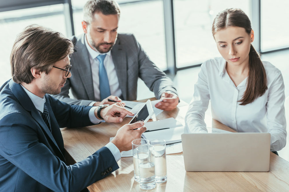 Three business professionals are seated at a wooden table in a bright office setting. A man in a suit is using a smartphone, while another man in a gray suit is focused on a tablet. A woman in a white shirt is working on a laptop. There are glasses of water on the table.