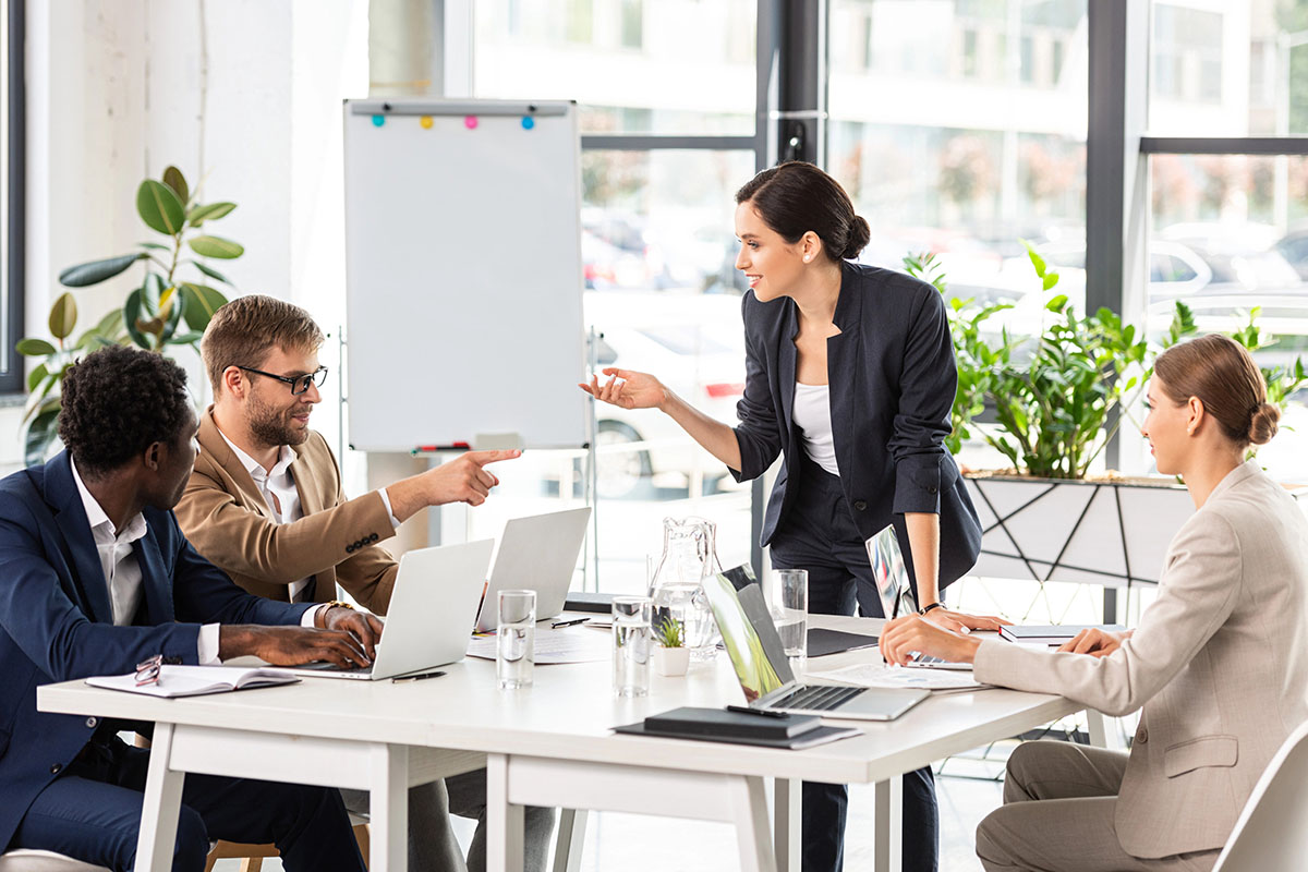 A diverse group of four professionals engaged in a meeting at a bright, modern office. A woman in a dark suit stands while pointing, engaging two seated male colleagues in conversation; one is wearing glasses and a light-colored suit. A woman in a light suit observes from the other side of the table, which holds laptops and glasses of water. A whiteboard is visible in the background, along with indoor plants.