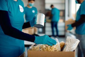 A volunteer wearing turquoise gloves and a shirt holds a can while placing food items, including pasta and beans, into a cardboard box. Other masked volunteers can be seen in the background, also working with food donations.