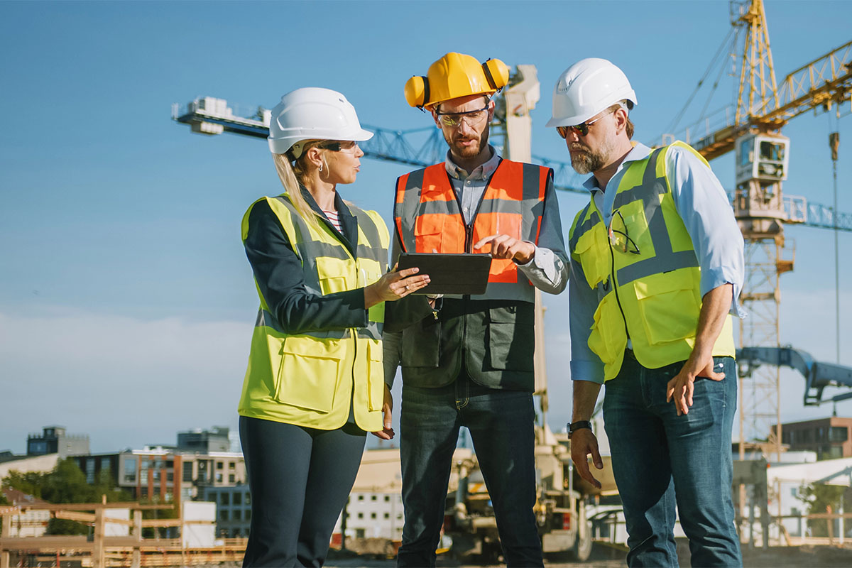 Three construction professionals, wearing safety helmets and reflective vests, discuss plans on a tablet at a construction site, with cranes and buildings in the background.