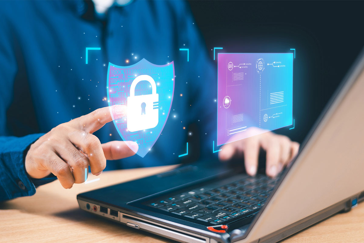 A person’s hand is reaching toward a digital shield icon with a padlock, representing cybersecurity, while using a laptop. The background features glowing data elements and abstract graphics related to connectivity.