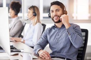 A young man with a beard, wearing a blue shirt and headset, smiles while speaking into a microphone at a call center, with two colleagues in the background.