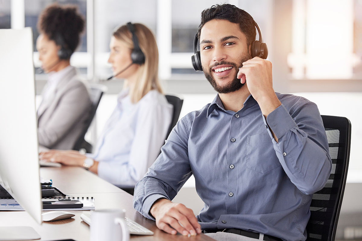 A young man with a beard, wearing a blue shirt and headset, smiles while speaking into a microphone at a call center, with two colleagues in the background.