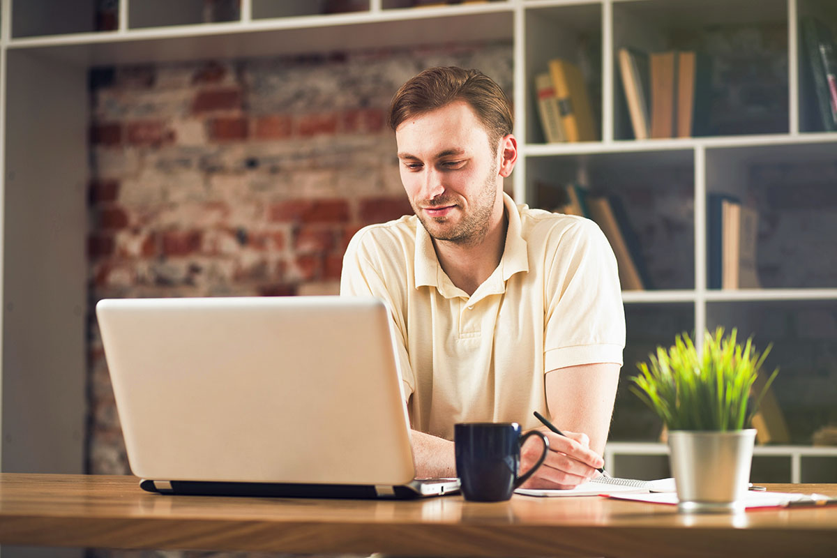 A young man in a light-colored polo shirt sits at a wooden desk, working on a laptop. He appears focused and is holding a pen while looking at the screen. A blue mug and a small potted plant are on the desk, with shelves of books in the background.
